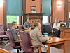 People seated and standing in a courtroom with wooden furnishings and tall windows.