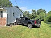 Black pickup truck parked beside a house with white siding and brick, on grass near trees.