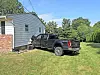 Black pickup truck parked beside a house with white siding and brick, on grass near trees.