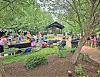People gathered around picnic tables near a pavilion in a park surrounded by trees and shrubs.