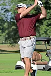 Golfer in a maroon shirt and gray shorts finishing a swing on a golf course near a golf cart.