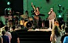 A live band performs on stage under green lighting with an audience standing in front.