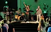 A live band performs on stage under green lighting with an audience standing in front.