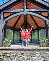 Two people pose with raised arms in front of a wooden pavilion with glass walls and potted plants.
