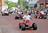 Several people drive go-karts down a brick street past crowds sitting and standing along the sidewalk.