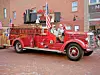A red vintage fire truck with two seated occupants is parked on a brick street beside a brick building.