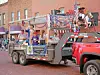 Una carroza decorada con gente y banderas es remolcada por una camioneta roja por una calle.