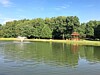 A lake with a water fountain, grassy bank, pavilion, and trees under a blue sky.