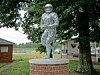 Stone statue of a football player on a brick pedestal in a park near trees and buildings.