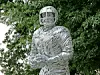 Silver metal sculpture of a human figure standing in front of green trees.