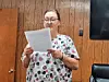 Woman reading from a sheet of paper in a wood-paneled room.