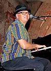 Man in a hat and striped shirt seated at a piano, singing into a microphone.