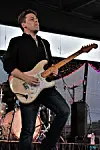 Guitarist playing a white electric guitar on stage with drums and speakers behind.