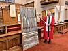 A clergy member in a red robe stands beside a large woven textile hanging in a church interior.