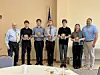 Seven people stand in a room holding plaques and awards with a U.S. flag behind them.