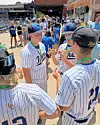 Youth baseball players in white pinstriped uniforms gather near the dugout at a stadium.