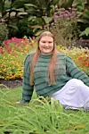 Woman sitting on grass in a garden with colorful flowers and green plants behind her.
