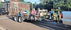 Workers ride on a flatbed trailer being towed along a street past houses and parked vehicles.