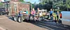 Workers ride on a flatbed trailer being towed along a street past houses and parked vehicles.