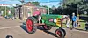 A green vintage tractor with red wheels passes people standing along a street during a community parade.