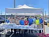 Group of volunteers standing behind a table of packaged items under a white canopy outdoors.