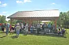 People gather under a pavilion with tables and chairs on a grassy field.