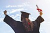 Graduate in cap and gown with arms raised, holding a diploma against the sky.