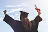 Graduate in cap and gown with arms raised, holding a diploma against the sky.
