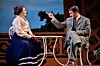 Two actors sit onstage at a small table, with one man gesturing toward a woman in a period costume.