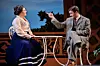Two actors sit onstage at a small table, with one man gesturing toward a woman in a period costume.