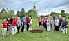 A group of people standing in a circle around a newly planted tree in a grassy outdoor setting.
