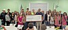 Large group posing with an oversized check in front of a green wall at an indoor event.