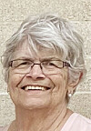 Smiling older woman with short gray hair, glasses, and a light top against a textured wall.