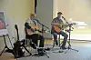 Two men seated on stools play acoustic guitars during an indoor performance.