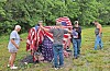 People stand on a grassy slope around a large American flag near trees.
