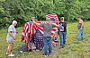 People stand on a grassy slope around a large American flag near trees.