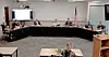 Officials seated around a U-shaped table in a meeting room with a podium and flags.