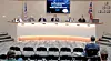 Council members seated behind a long curved desk in a meeting room with audience chairs in the foreground.