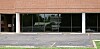 Vacant storefront with brick exterior, large glass windows, and an empty parking lot in front.