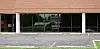 Vacant storefront with brick exterior, large glass windows, and an empty parking lot in front.