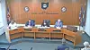 Officials seated at a curved dais in a city commission meeting room with flags and seals on the wall.