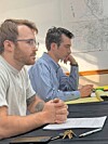 Two men seated at a table, looking at documents in a meeting room with a wall map behind them.