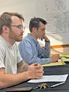 Two men seated at a table, looking at documents in a meeting room with a wall map behind them.