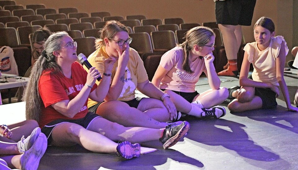 Several people sit cross-legged on a stage floor in front of rows of empty theater seats.
