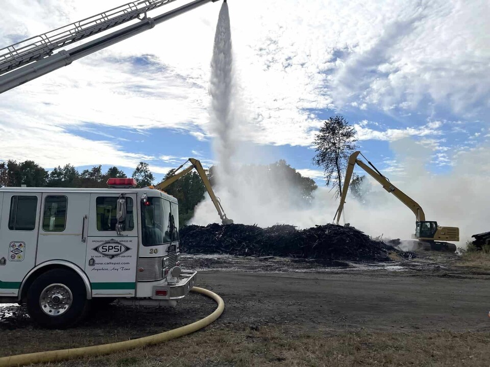 Emergency personnel use a ladder truck to spray water on a smoldering pile of railroad ties as excavators work from the outside to the inside of the pile to gain access to the center.